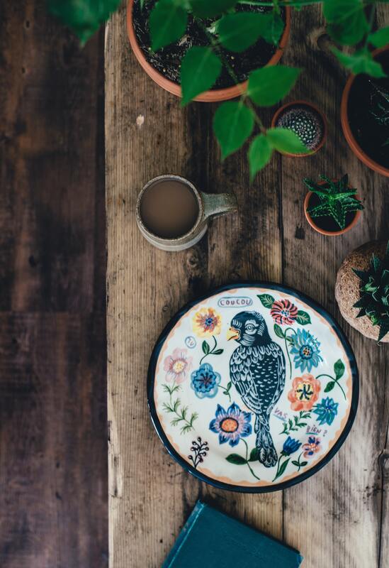 An embroidery hoop sits on a wooden table. It's surrounded by plants and a cup of coffee. Embroidered on the hoop is a blue bird.
