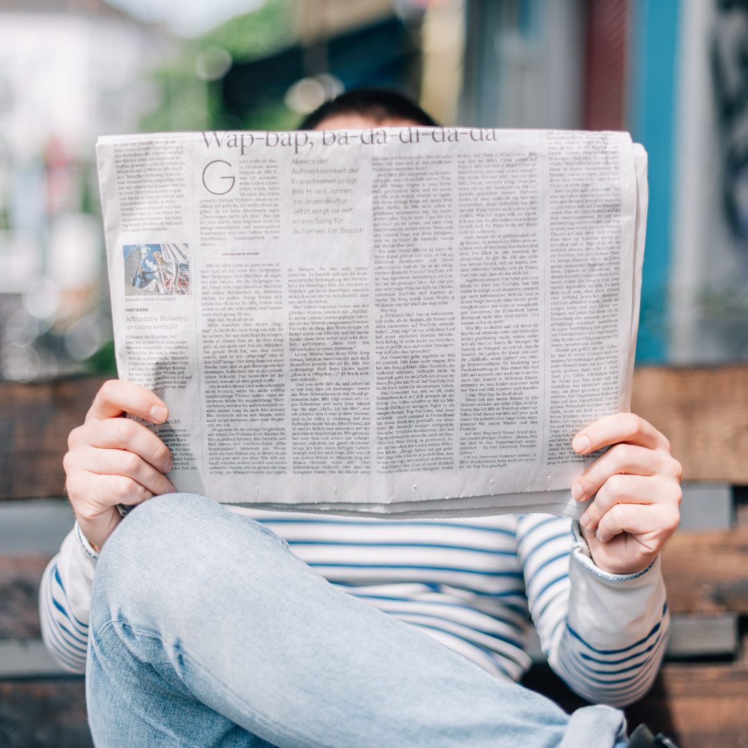 A person sits on a bench reading a newspaper. Their face is covered by the paper.