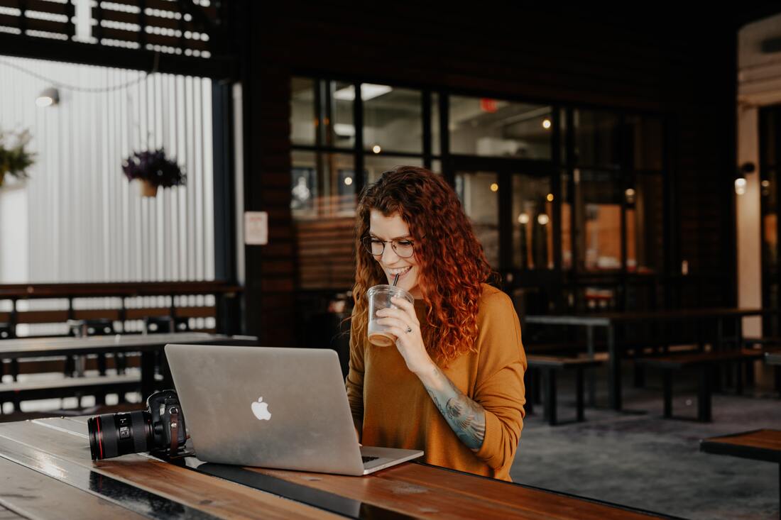 A woman wearing a yellow sweater types on an Apple Macbook. She is sitting alone at a table, sipping on an iced latte.
