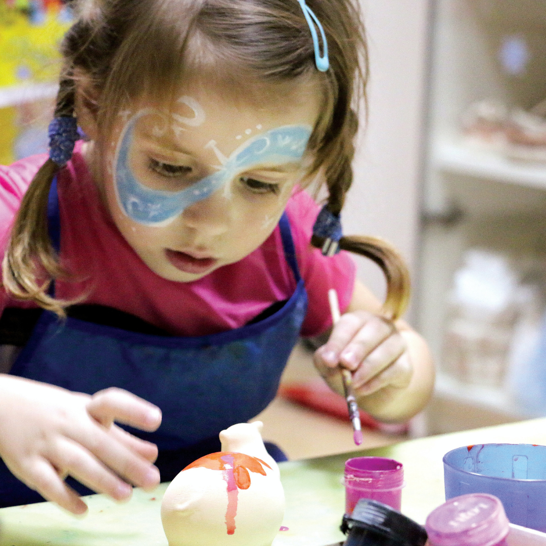 A young girl is painting a ceramic lamb. Her face is painted with blue swirls.