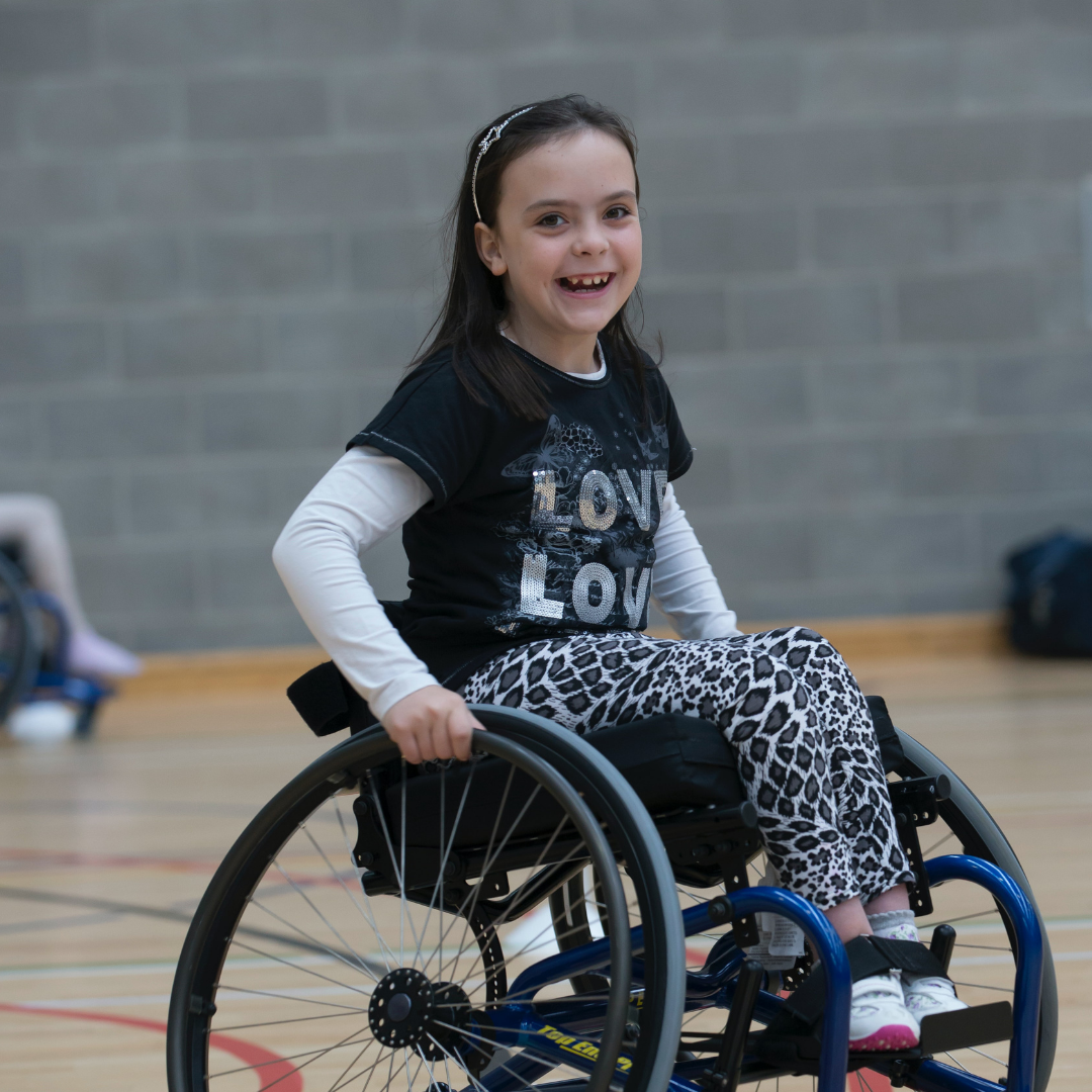 A young girl plays basketball. She is using a wheelchair. Her hair is pulled back by a headband and she's smiling broadly.
