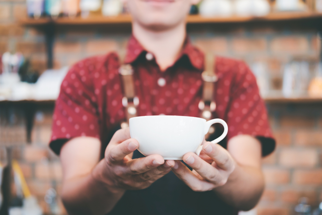 A barista wearing a red shirt and denim apron is holding a coffee mug.